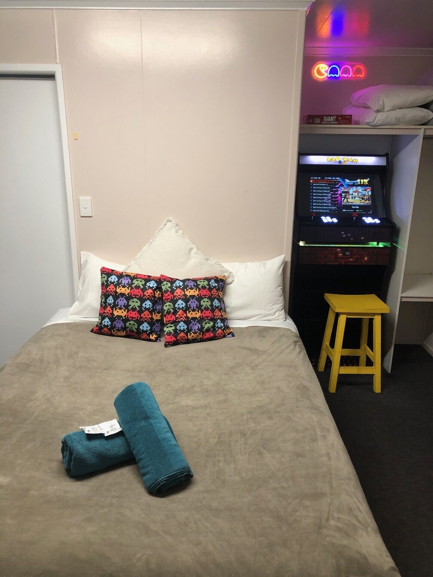 Une chambre d'hôtel avec un lit, une borne d'arcade lumineuse et un petit tabouret.