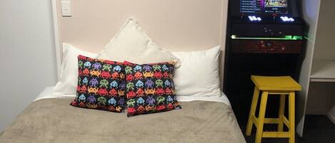 Una habitación de hotel con una cama, una máquina de juegos de neón y un taburete pequeño.
