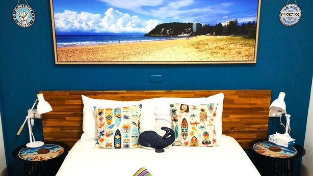 Habitación de hotel con una cama grande, dos mesitas de noche, una mesilla auxiliar, un televisor montado en la pared y una pintura con vista al mar.