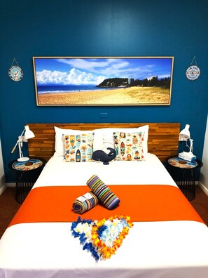 A hotel room with a large bed, two bedside tables, a nightstand, a wall-mounted TV, and a beach view painting. - Acacia Motel (Chinchilla)