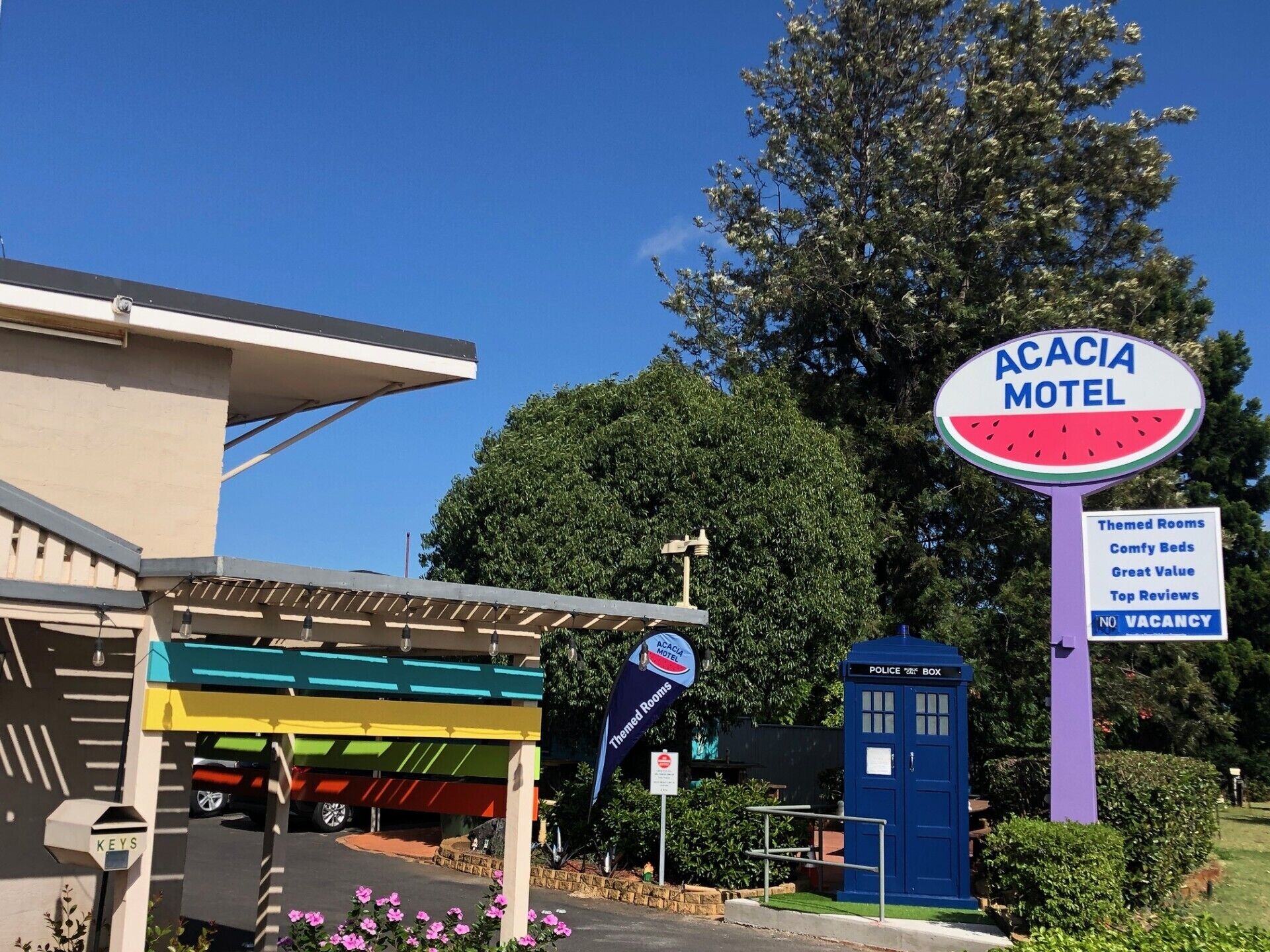 Um motel com placa anunciando quartos temáticos, camas confortáveis, ótimo custo-benefício e avaliações excelentes. Há uma caixa de polícia e uma placa indicando quartos indisponíveis.