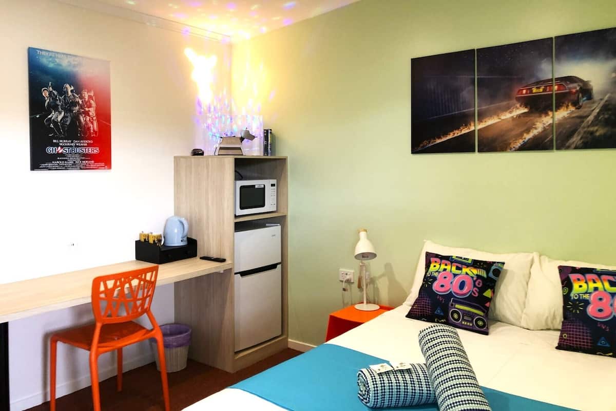A bedroom with a bed, a desk, a chair, a microwave, a small refrigerator, and a ceiling light fixture.