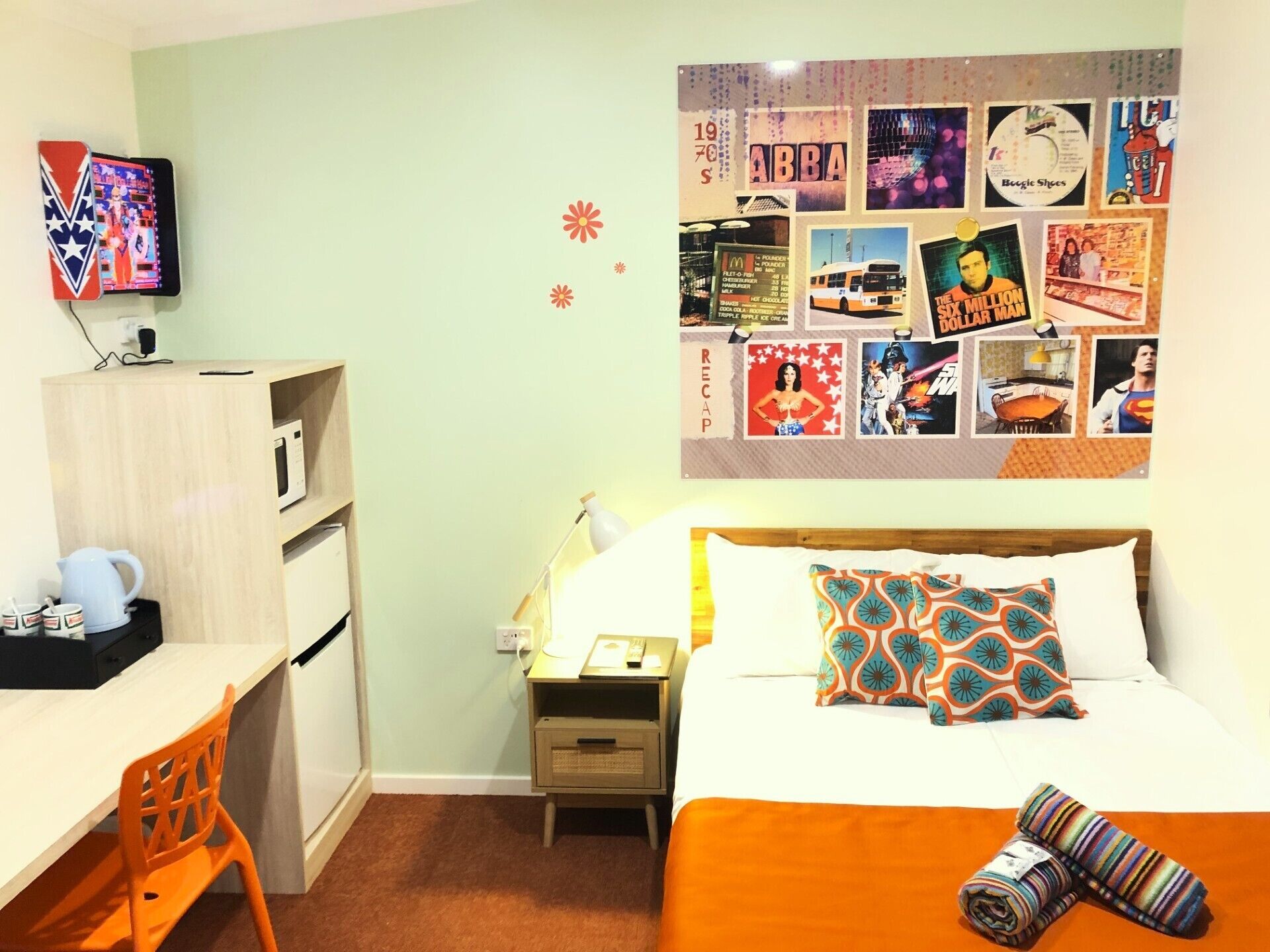 Een hotelkamer met een bed, een bureau, een stoel, een nachtkastje, een lamp, en een wand versierd met verschillende posters en foto's.
