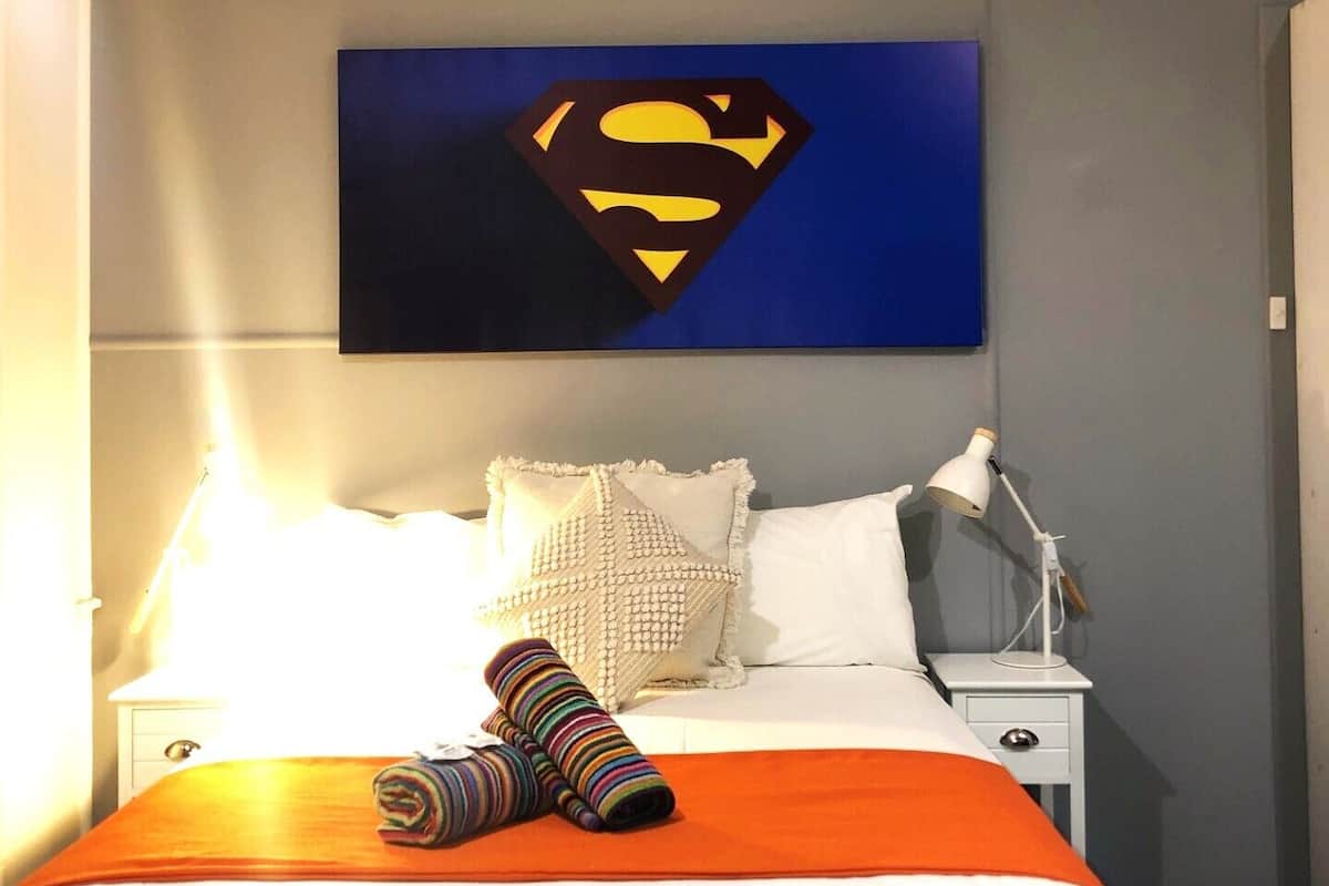 A hotel room with a bed, a nightstand, a lamp, and a Superman logo on the wall.