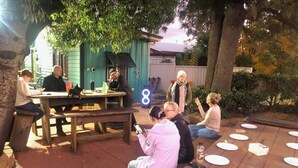 Un grupo de personas sentadas en mesas de picnic de madera al aire libre, con un edificio verde al fondo.