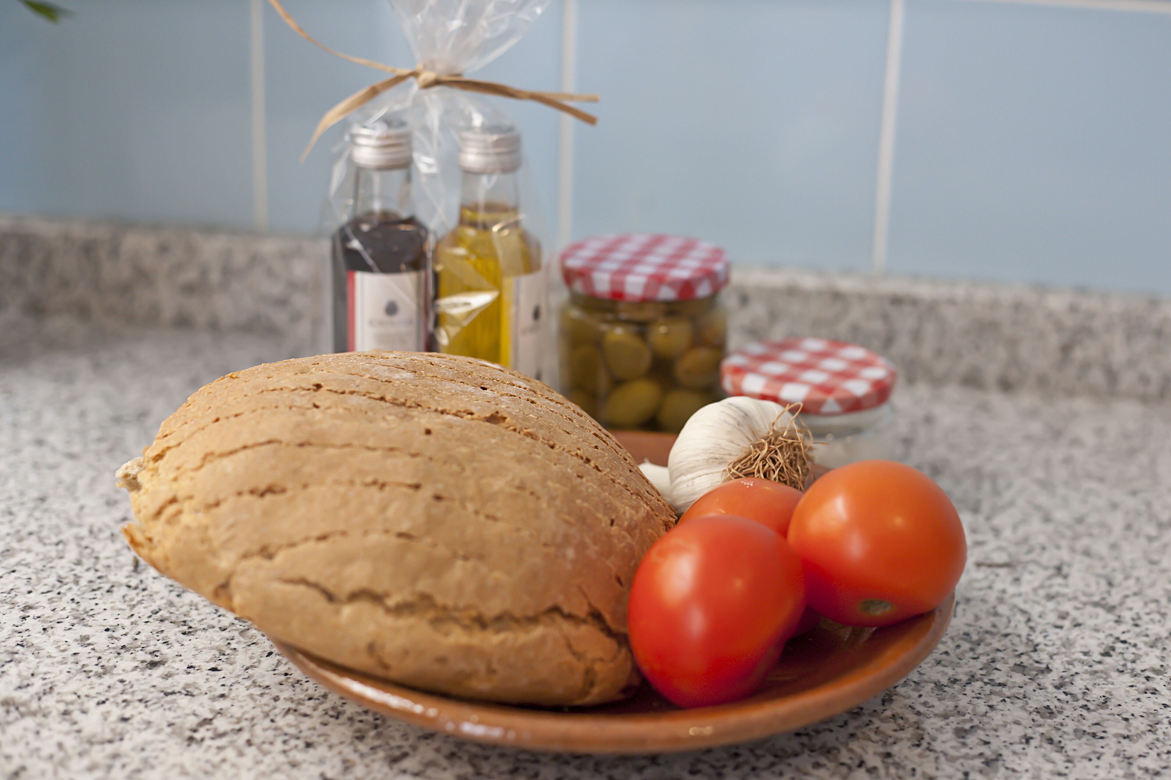 A loaf of bread, tomatoes, garlic, and jars of olive oil on a kitchen counter.
