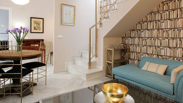 A modern living room with a round dining table, a blue sofa, a staircase, and a bookshelf filled with books. - Celinda Terrace. 2 bedrooms, 2 bathrooms, private terrace (Seville)
