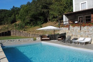 Une piscine entourée de chaises longues et d’un parasol, le tout bordé de murs en pierre et d’une clôture en bois.
