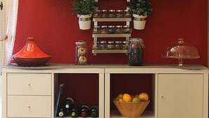 Une cuisine avec un mur rouge, des étagères remplies d’ustensiles de cuisine, un panier de fruits et des paniers suspendus contenant des plantes.