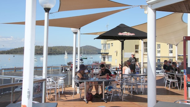 An outdoor dining area with tables and chairs, a view of the water, and a building in the background.