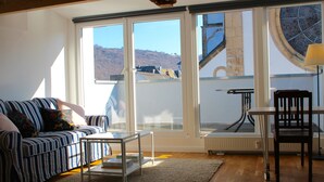 Una sala de estar con un sofá, una mesa de centro y una silla. Hay ventanas amplias con vistas a un edificio y una colina.