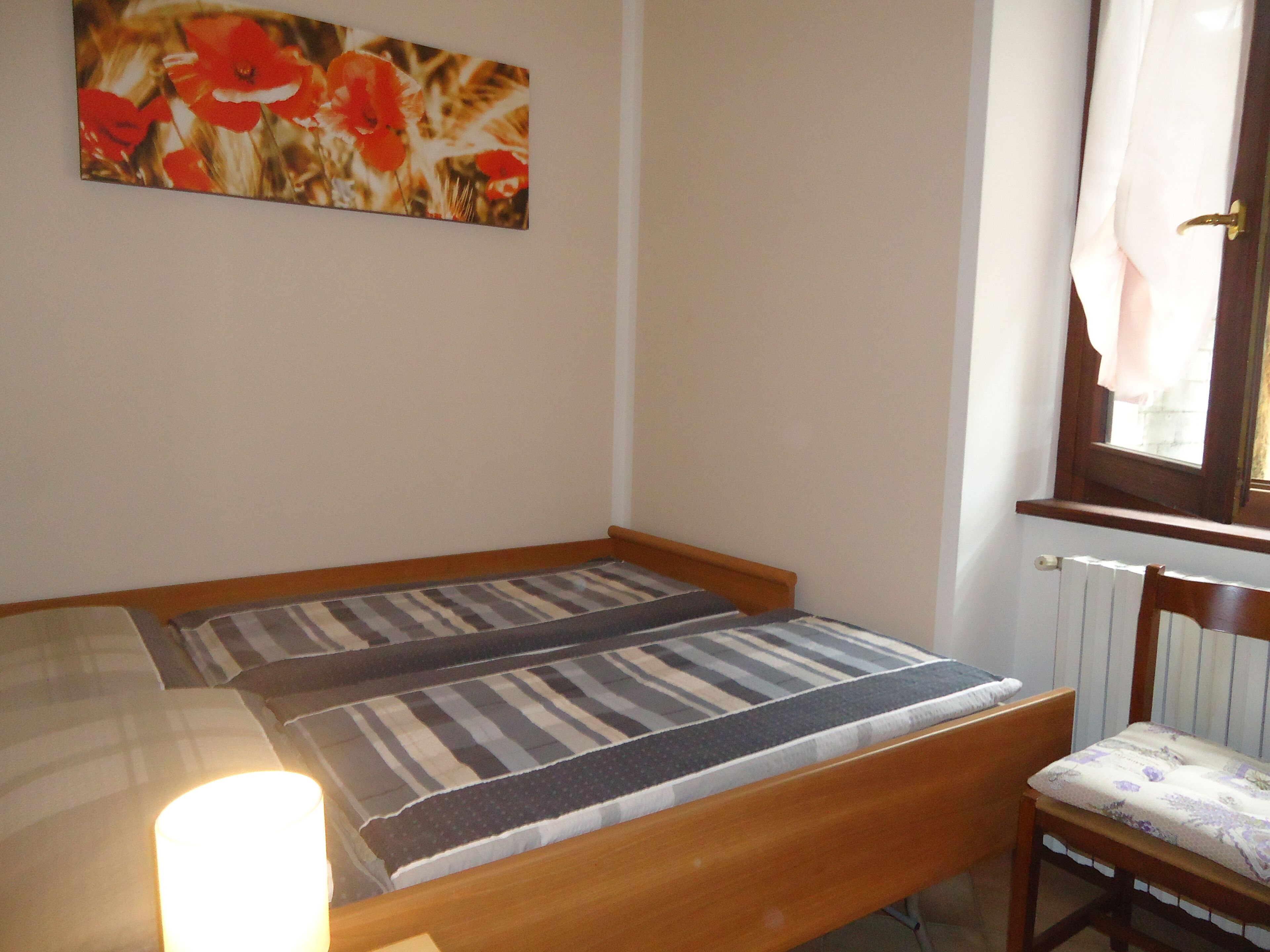 Une petite chambre avec un lit simple, un cadre en bois, une literie à rayures, une chaise avec un coussin à motifs floraux et une fenêtre avec des rideaux.
