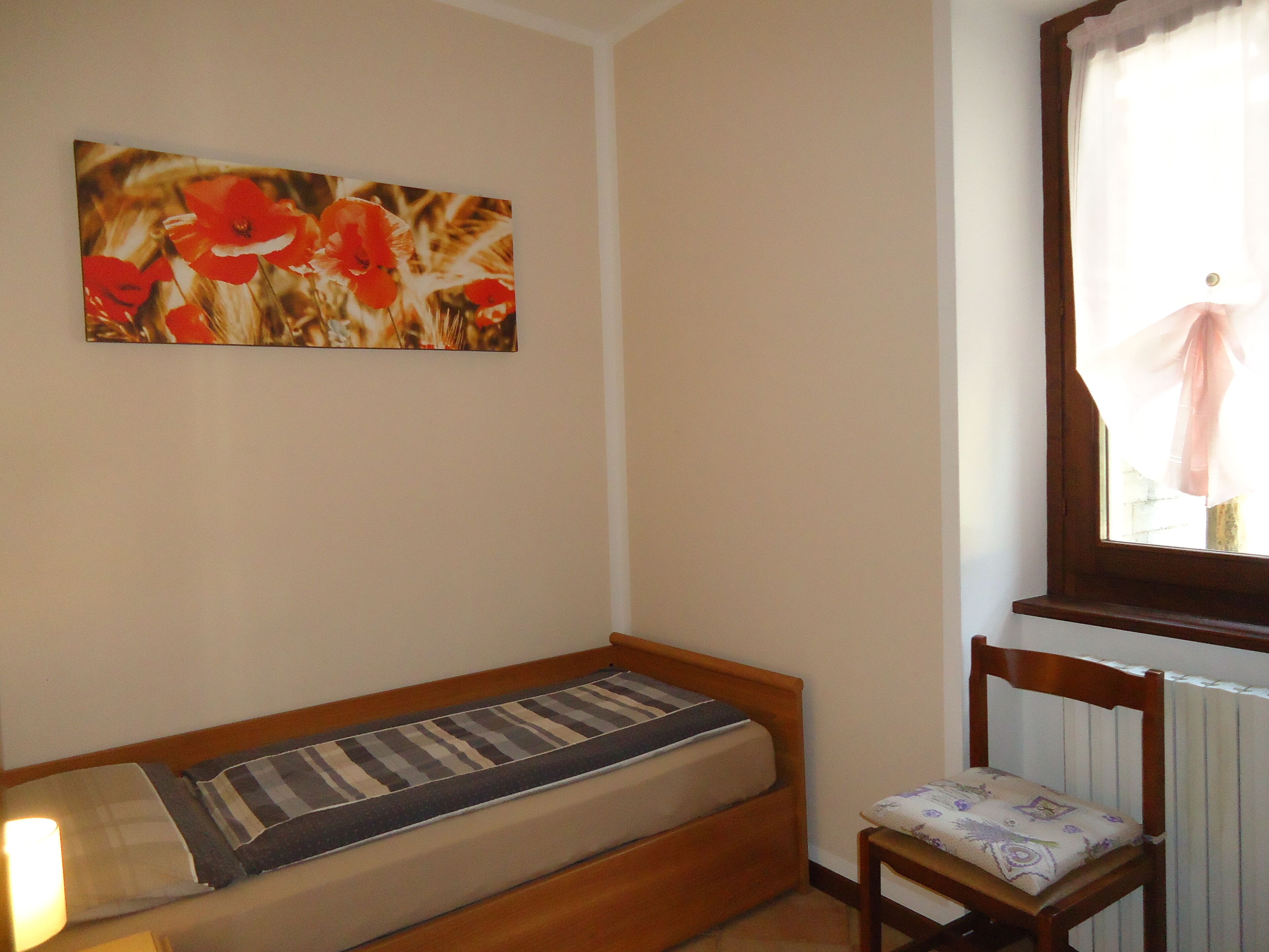 A small, single-bed room with a wooden headboard, a bedside table with a lamp, and a chair with a cushion.