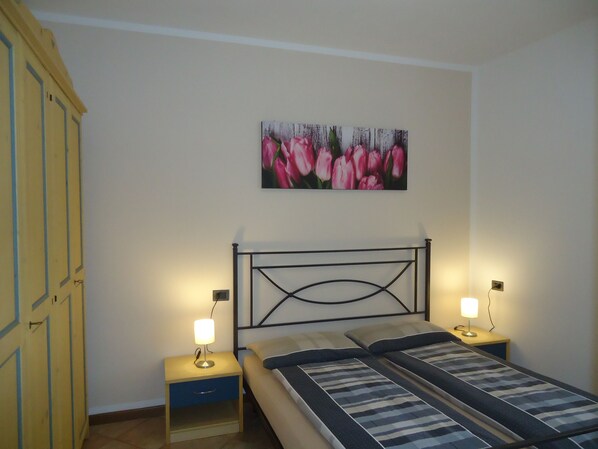 A bedroom with a bed, two bedside tables with lamps, a nightstand, a wardrobe, and a painting of pink tulips on the wall.