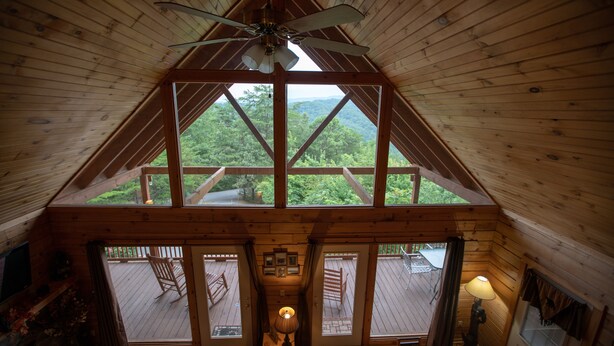 Une cabane en bois avec un toit en pente, un ventilateur de plafond, une cheminée et une vue sur la forêt environnante.