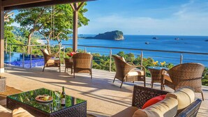 Una terraza con mobiliario de mimbre, una mesa de vidrio y vistas al mar y una pequeña isla.