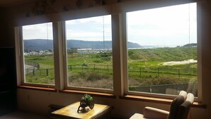 Een kamer met drie grote ramen met uitzicht op een kustlandschap, inclusief een weg en heuvels in de verte.