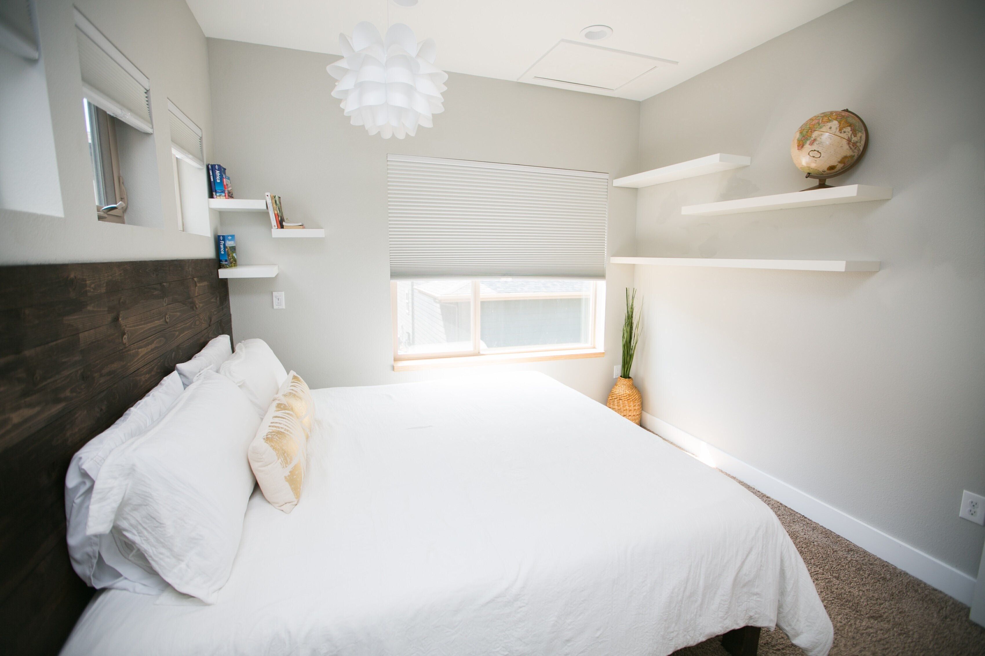 Un dormitorio con cama, paredes blancas, una ventana con persianas, cabecera de madera y un globo terráqueo en un estante.