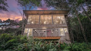 Una cabaña de madera de dos pisos con ventanas amplias y un balcón, rodeada de un bosque denso.