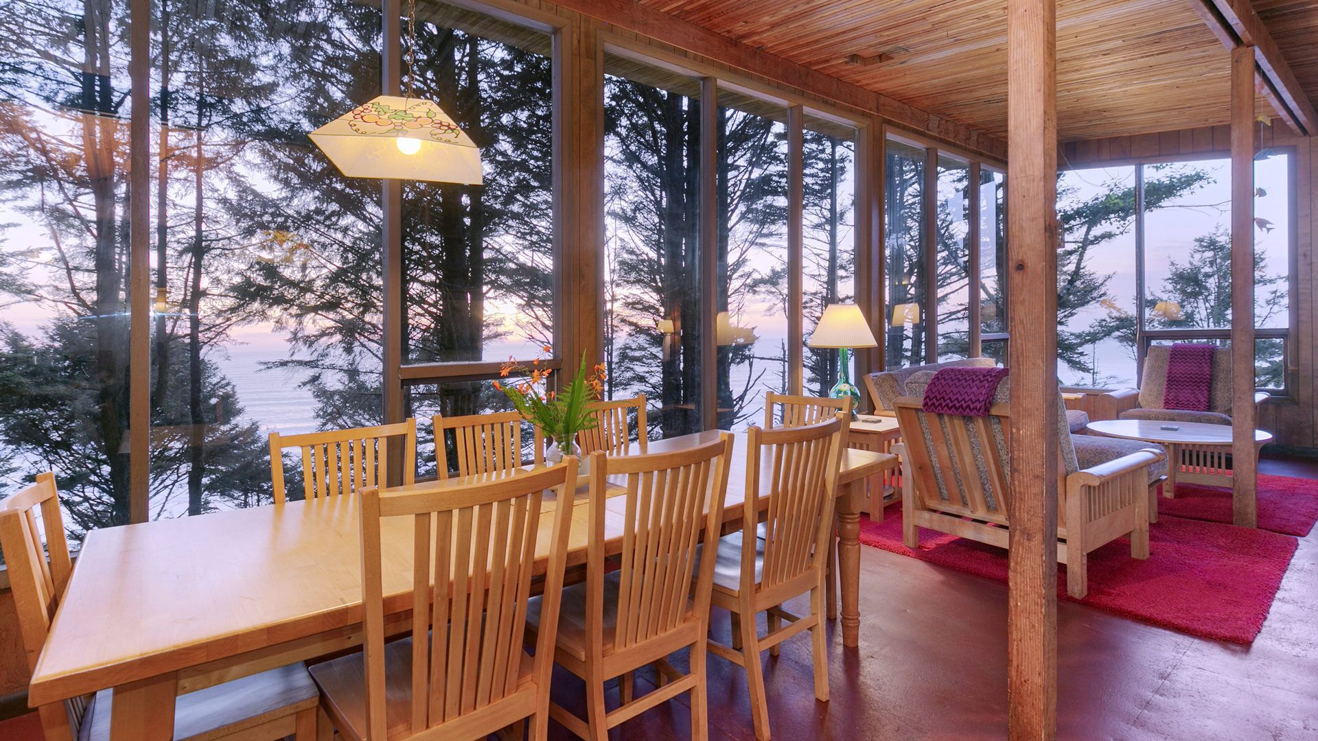 Un coin repas en bois, avec une table et des chaises, de grandes fenêtres et une vue sur les arbres et l’eau.