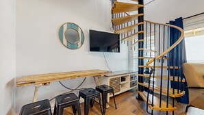 A modern living space with a wooden desk, black stools, a TV mounted on the wall, and a spiral staircase. - Beachside Joy 1 Bedroom Townhome Pet friendly (Panama City Beach)