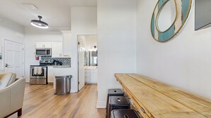 A modern kitchen with white cabinets, a microwave, and a stove. There is a wooden bench and a trash can in the living area. - Beachside Joy 1 Bedroom Townhome Pet friendly (Panama City Beach)