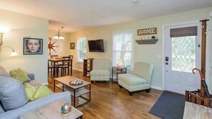 Un salon comprenant un canapé, des fauteuils, une table basse, une table à manger avec des chaises, un téléviseur et une porte sur laquelle est apposé un panneau indiquant « MAISON ».