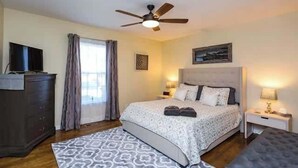 Une chambre à coucher comprenant un lit, un ventilateur de plafond, une commode, un téléviseur et une fenêtre avec des rideaux.