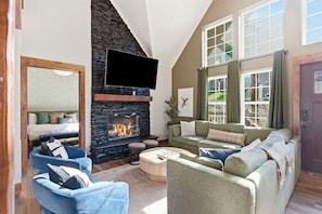 A living room with a stone fireplace, a flat-screen TV, and a large window with curtains.