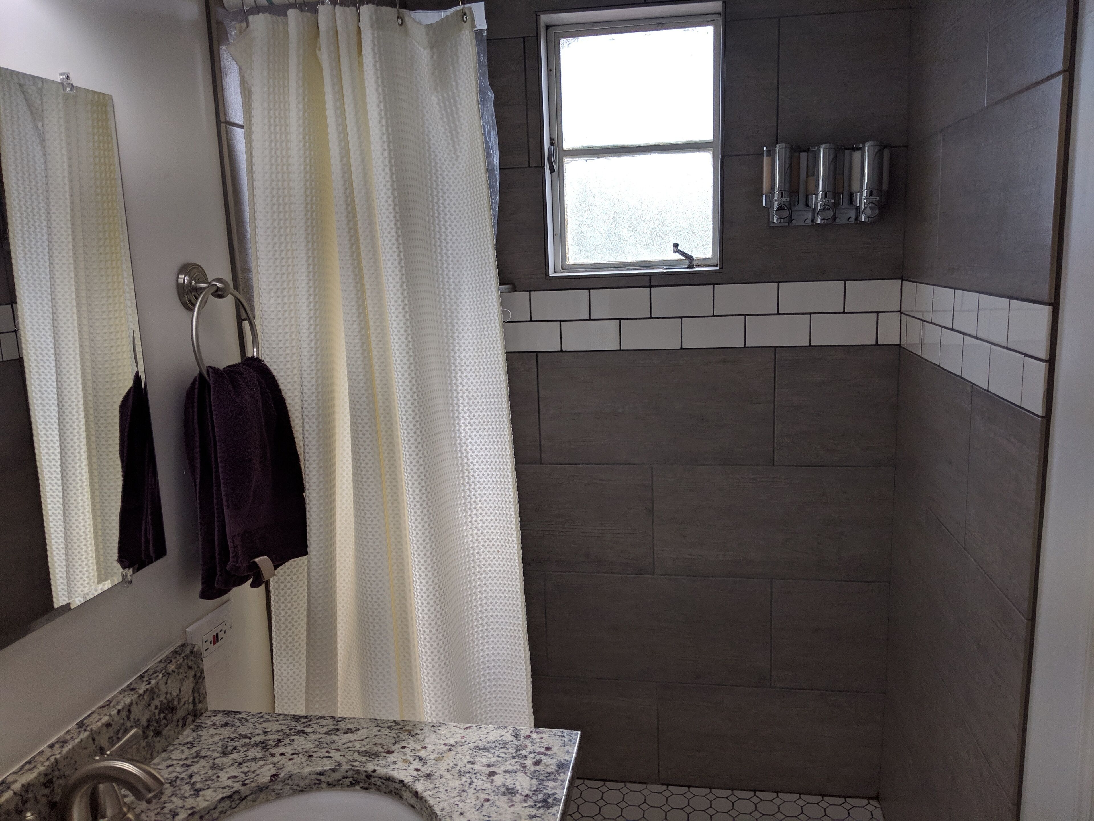 A bathroom with a shower, a sink with a marble countertop, a mirror, and a window.