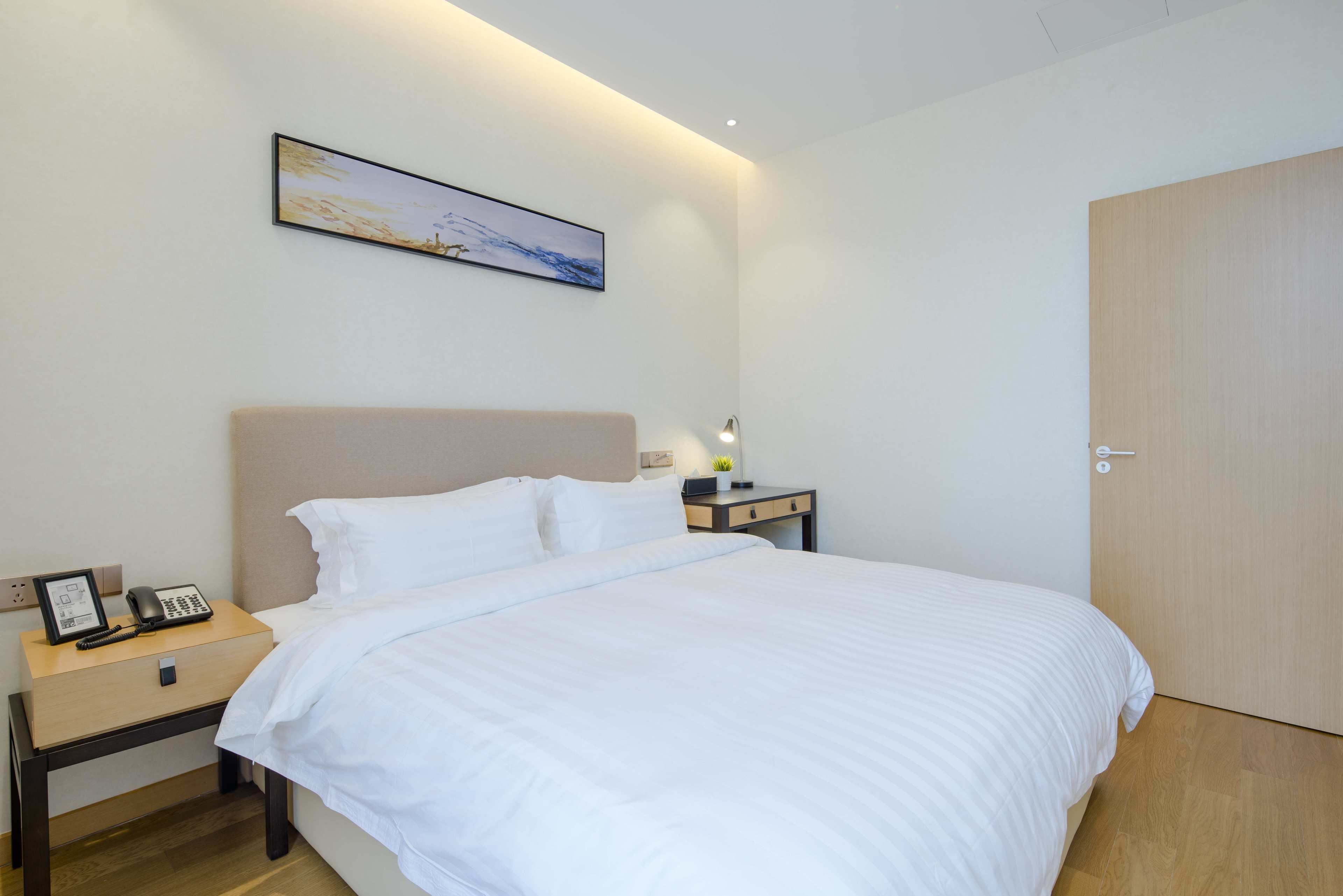 A neatly made bed in a hotel room with a headboard, a bedside table with a lamp, a phone, and a picture frame.