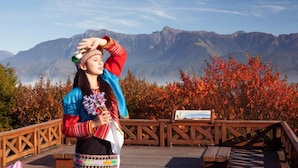 Une personne vêtue d’un costume traditionnel se tient sur une terrasse en bois, avec une chaîne de montagnes en arrière-plan.