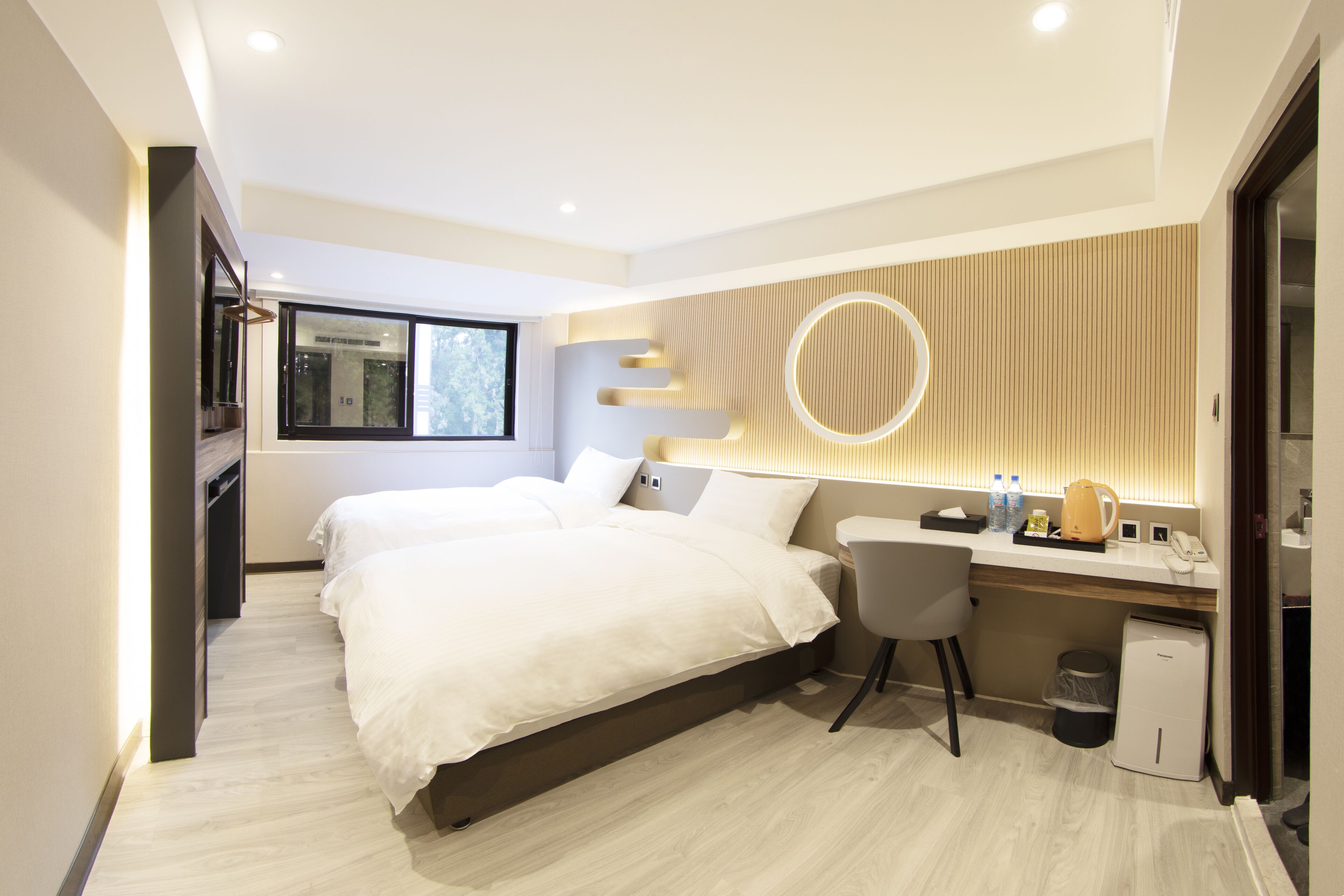 A modern hotel room with two beds, a desk, a chair, a small trash bin, and a circular light fixture on the wall.