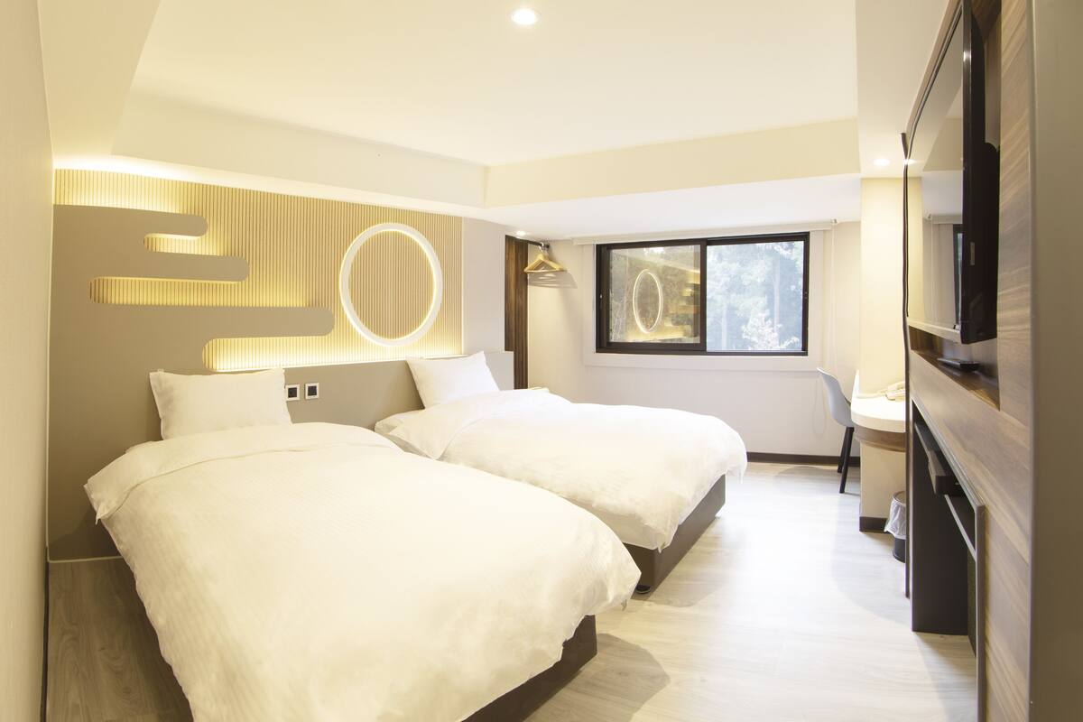 Une chambre d’hôtel moderne avec deux lits, un grand miroir, une télévision et une fenêtre donnant sur des arbres.