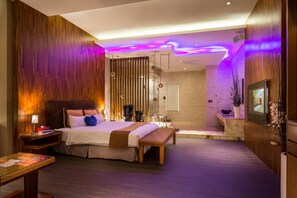 Ein modernes Hotelzimmer mit einem großen Bett, Nachttischen, einer Bank, einem Fernseher und einem Badezimmer mit Badewanne und Dusche.