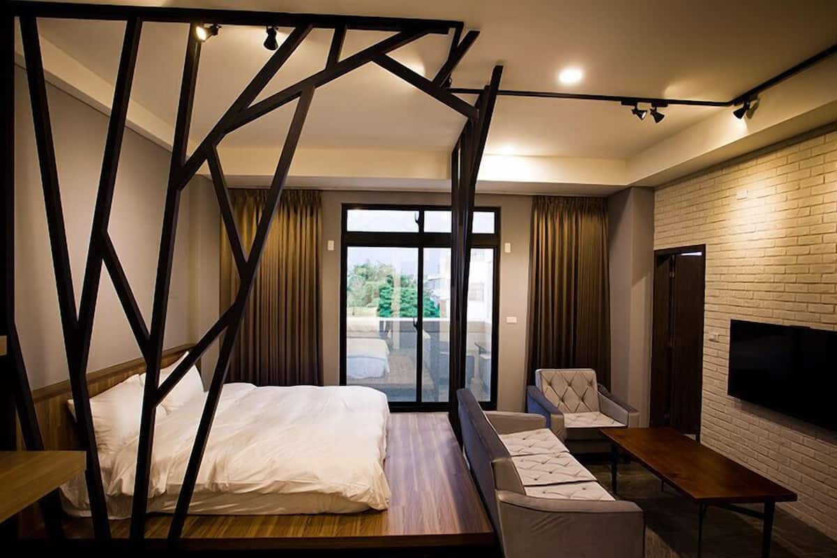Habitación de hotel moderna con una cama grande, zona de estar, televisor y balcón con vistas a zonas verdes.