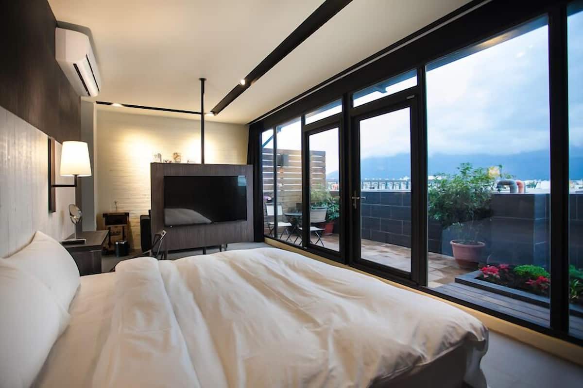 Habitación de hotel moderna con una cama grande, un televisor de pantalla plana, un escritorio y un balcón con plantas en macetas.