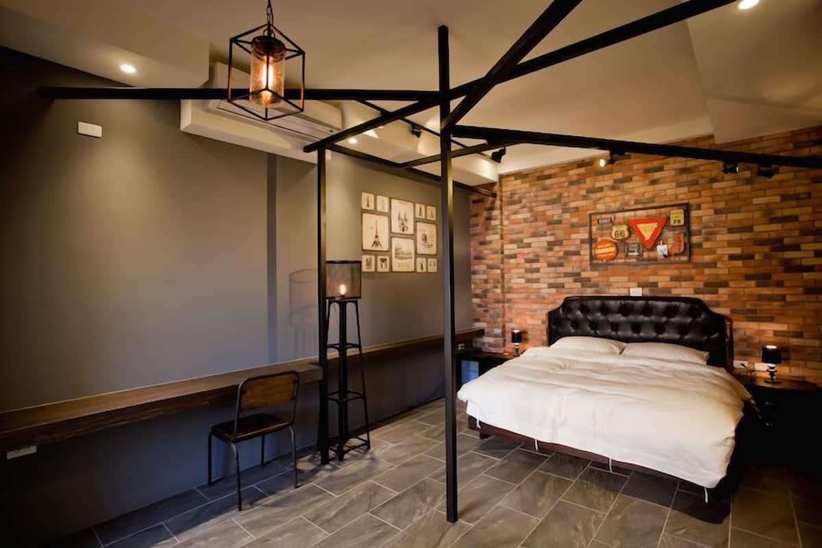 Un dormitorio con pared de ladrillo, una cama, una mesita de noche, una silla, un taburete y una lámpara colgante.