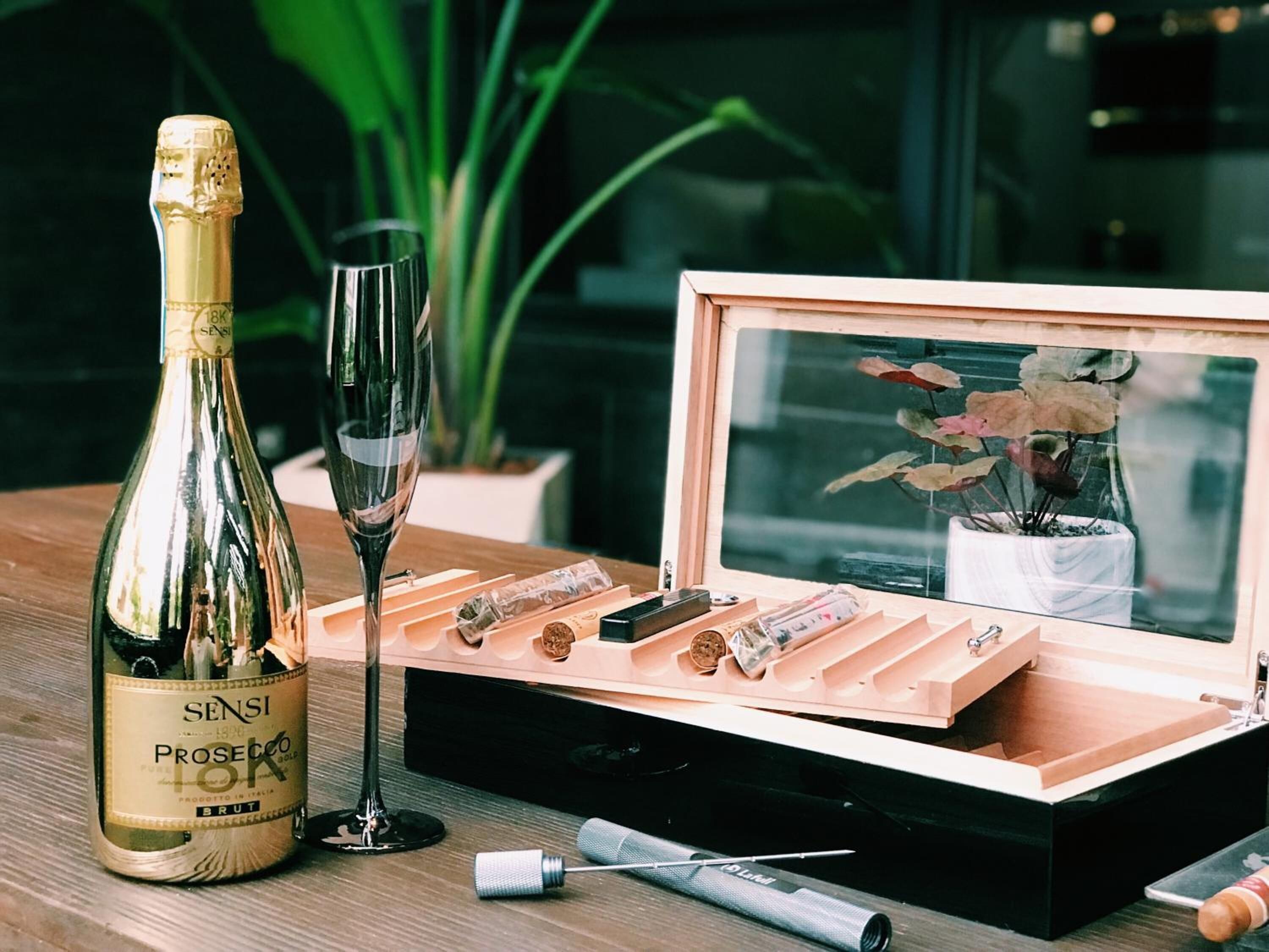 A bottle of Sensi Prosecco, a wine glass, and a wooden box with a corkscrew and other items on a wooden table.