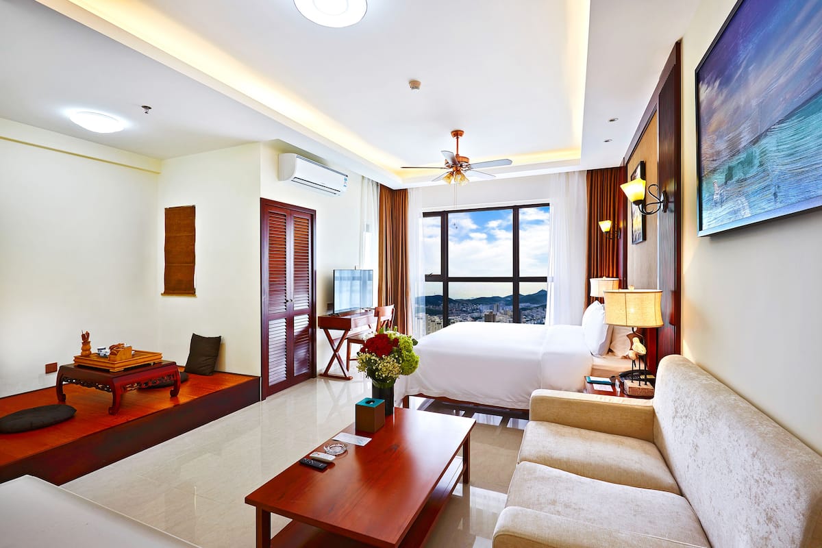 Habitación de hotel con cama, sofá, mesita de centro, escritorio y un amplio ventanal con vistas a la ciudad.