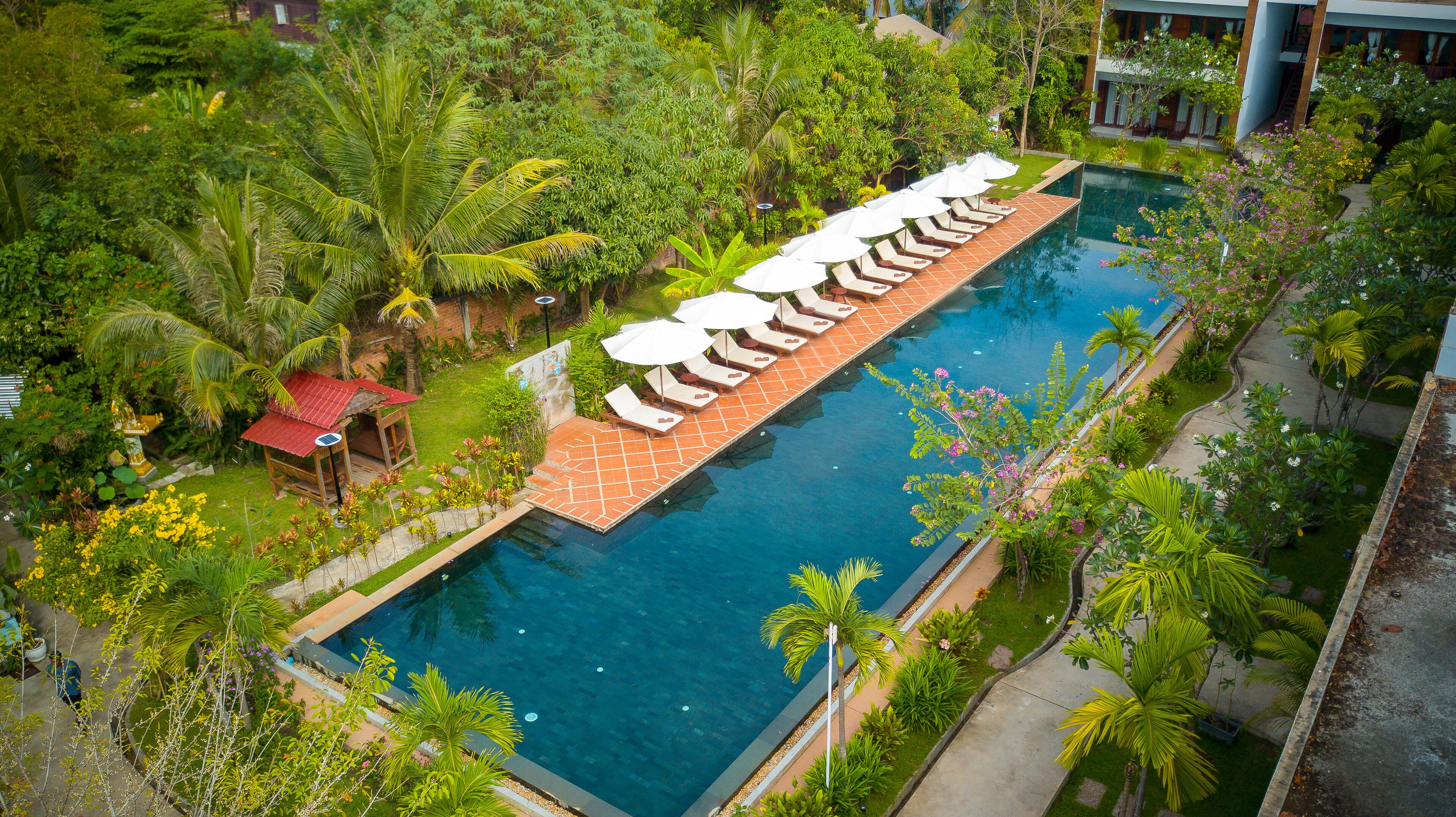 Une piscine rectangulaire dotée d’une terrasse carrelée, entourée d’une végétation luxuriante et de palmiers.