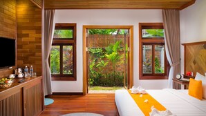 A hotel room with a bed, a desk with a TV, and a view of a garden through a door. - Khmer House Resort (Siem Reap)