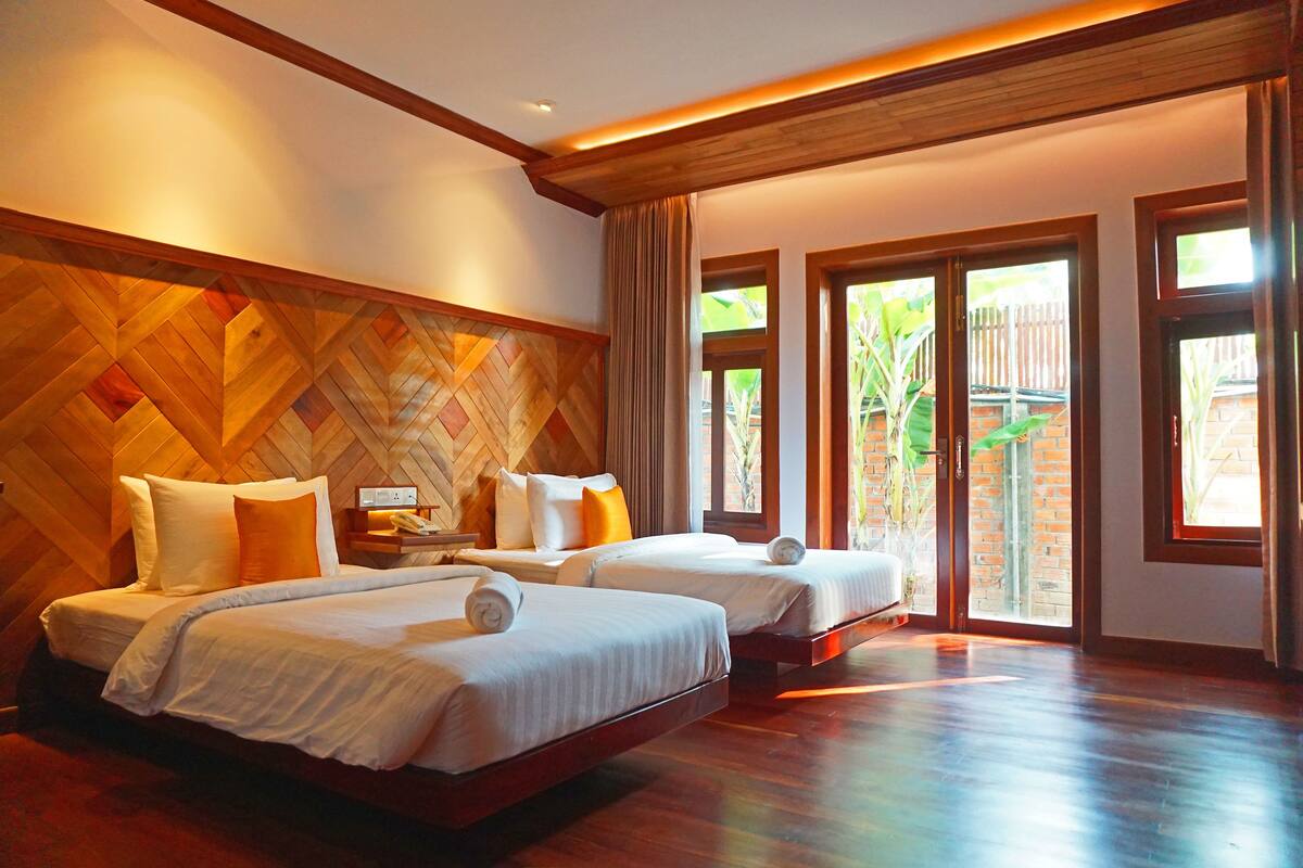 Une chambre d’hôtel avec deux lits, un plancher en bois, un mur orné d’un motif en chevrons et de grandes fenêtres donnant sur un paysage verdoyant.