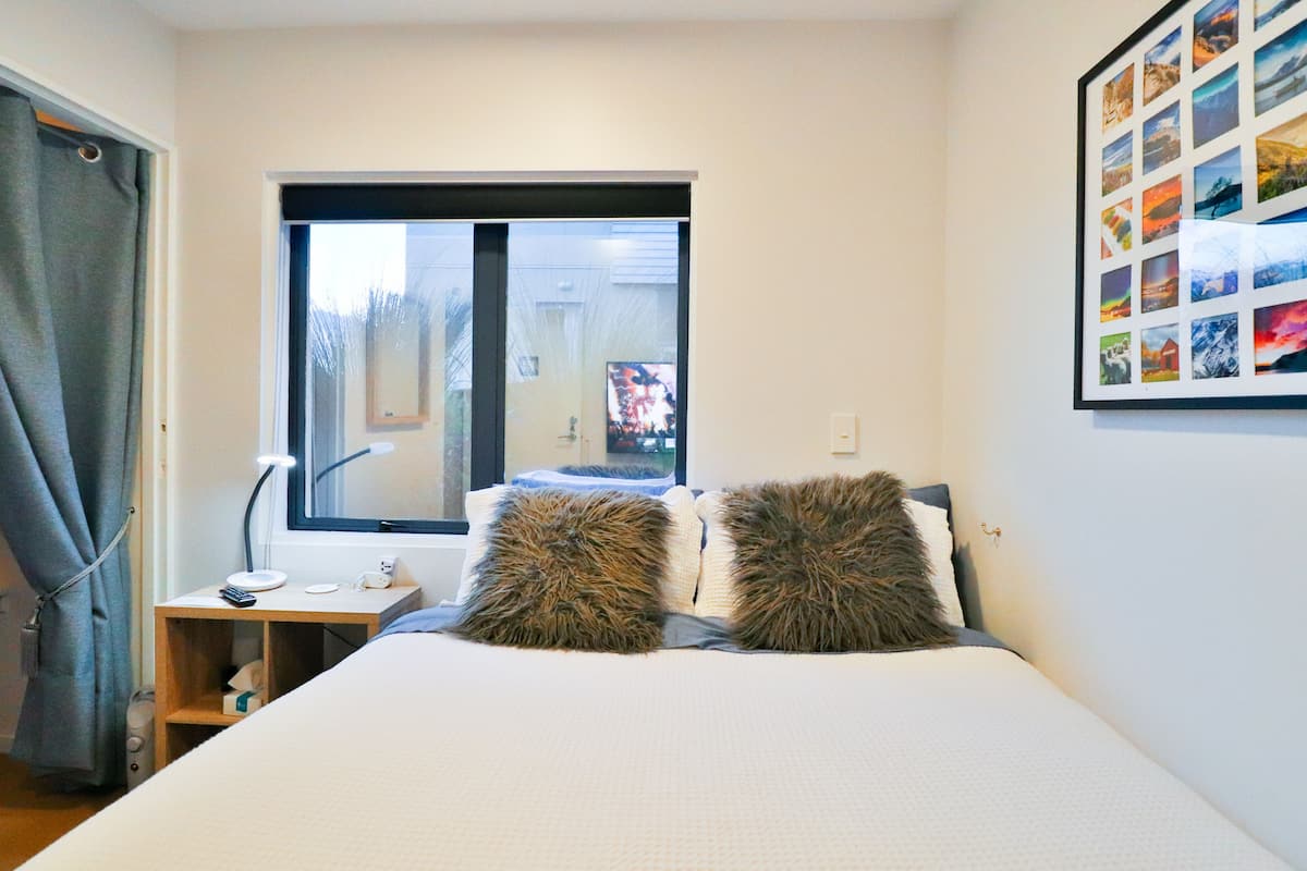 A bedroom with a bed, two fluffy pillows, a bedside table with a lamp, a window, and a wall-mounted photo collage.