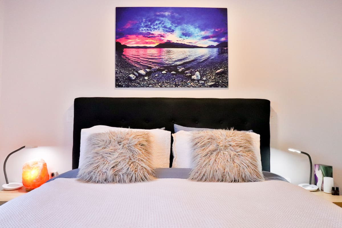 A bedroom with a bed, two fluffy pillows, a nightstand with a lamp, and a wall-mounted painting of a sunset over water.