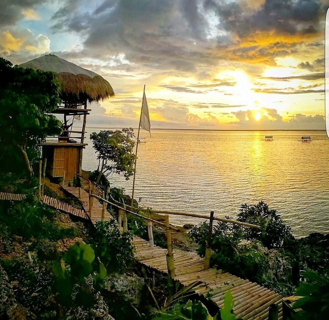 A wooden deck leading to a thatched-roof structure overlooking a calm sea at sunset.