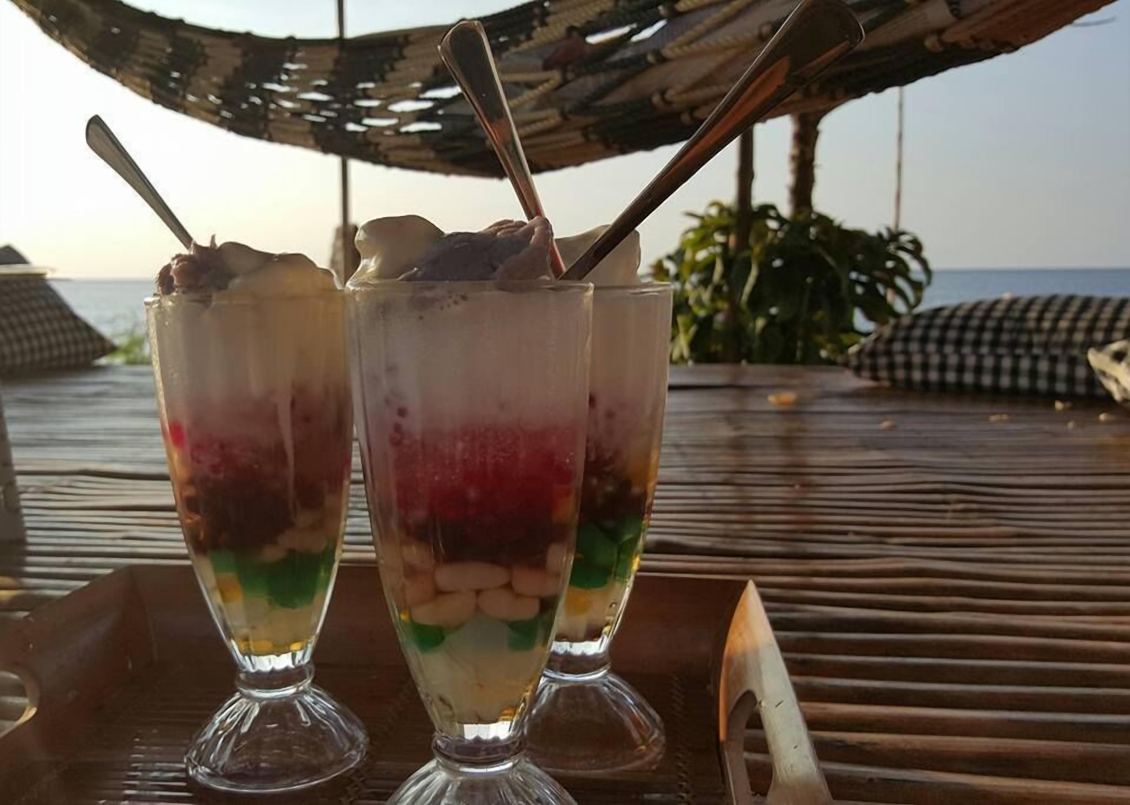 Dos postres en capas sobre una bandeja de madera, junto a una cuchara metálica, sobre una mesa de madera al aire libre, con un paraguas tejido y palmeras al fondo.