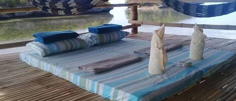 A hammock under a thatched roof, a bed with striped bedding, and a view of a calm body of water.