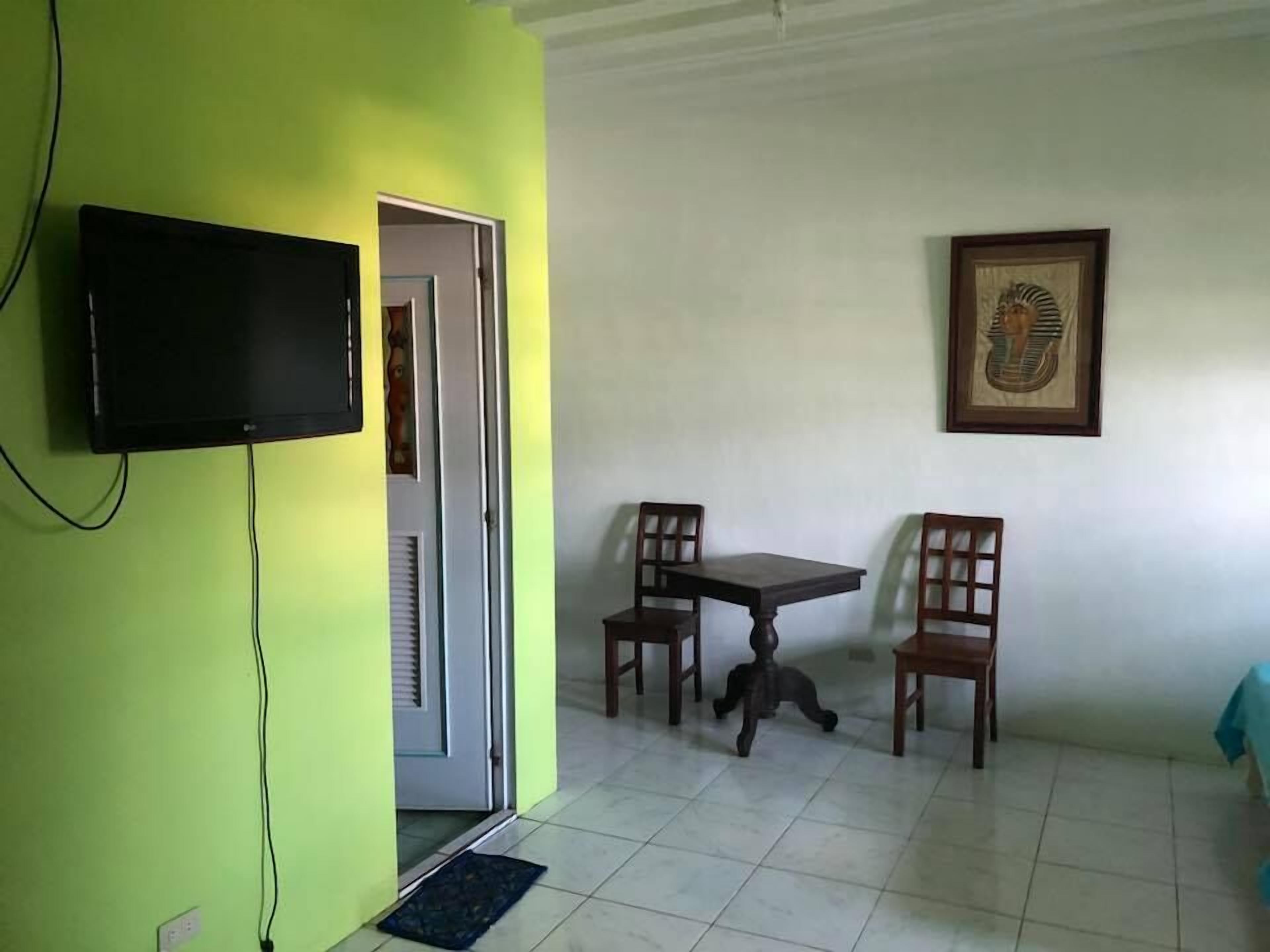 Habitación con televisión montada en una pared verde, puerta que da a otra habitación, una pequeña mesa con dos sillas y un cuadro enmarcado en la pared.