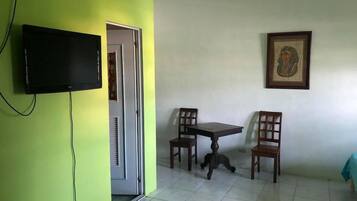 Habitación con televisión montada en una pared verde, puerta que da a otra habitación, una pequeña mesa con dos sillas y un cuadro enmarcado en la pared.